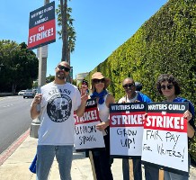 USA_ Streikposten der Drehbuchautor*innen - vier Menschen stehen mit Schildern vor einer grünen Hecke