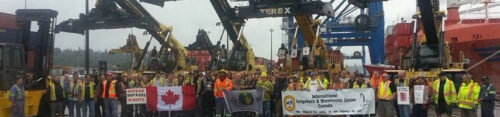 Dutzende Hafenarbeiter*innen Kanadas stehen in Vancouver vor den Kränen mit Transparenten